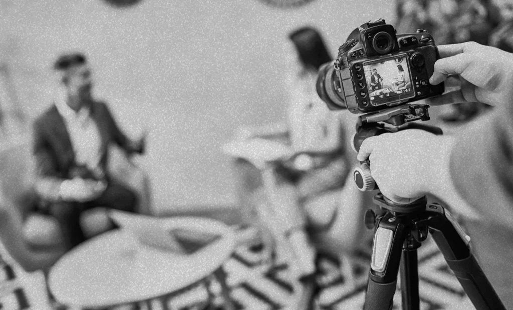 Man and woman sitting and talking, out of focus. Camera in the foreground recording their discussion.